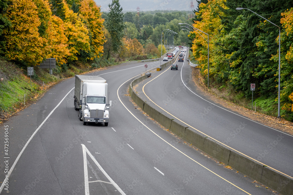 Industrial white big rig semi truck with tall cab transporting cargo in dry van semi trailer running for delivery on the wide divided highway road with autumn forest trees on the sides Industrial white big rig semi truck with tall cab transporting cargo in dry van semi trailer running for delivery on the wide divided highway road with autumn forest trees on the sides