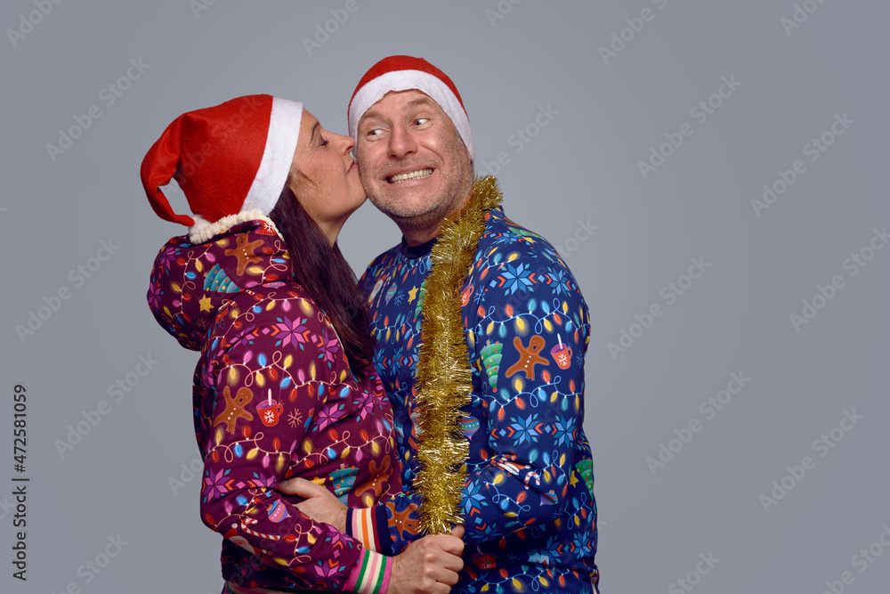 Affectionate middle-aged couple in festive holiday themed Christmas outfits embracing and kissing as they celebrate Xmas together over a grey background in side view Affectionate middle-aged couple in festive holiday themed Christmas outfits embracing and kissing as they celebrate Xmas together over a grey background in side view