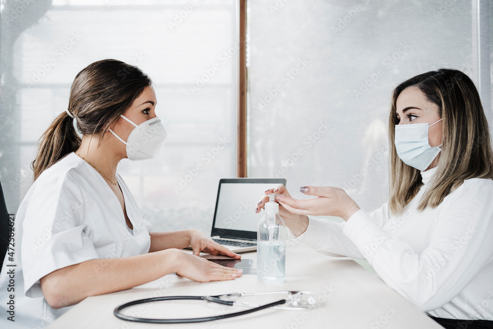 Patient taking sanitiser while talking with female doctor at desk in hospital during pandemic Patient taking sanitiser while talking with female doctor at desk in hospital during pandemic