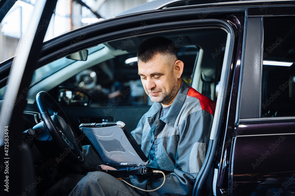 Smiling competent middle age serviceman making car diagnostics with laptop in a workshop. Auto service, repair, maintenance concept. Smiling competent middle age serviceman making car diagnostics with laptop in a workshop. Auto service, repair, maintenance concept.