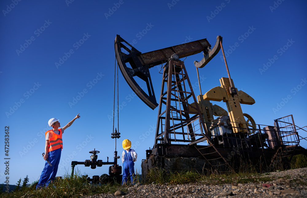Low view of mechanic showing process of petroleum extraction and pointing  finger up to working oil pump to little boy. Adult oil worker introducing  child to his job. Oil industry concept. Photos
