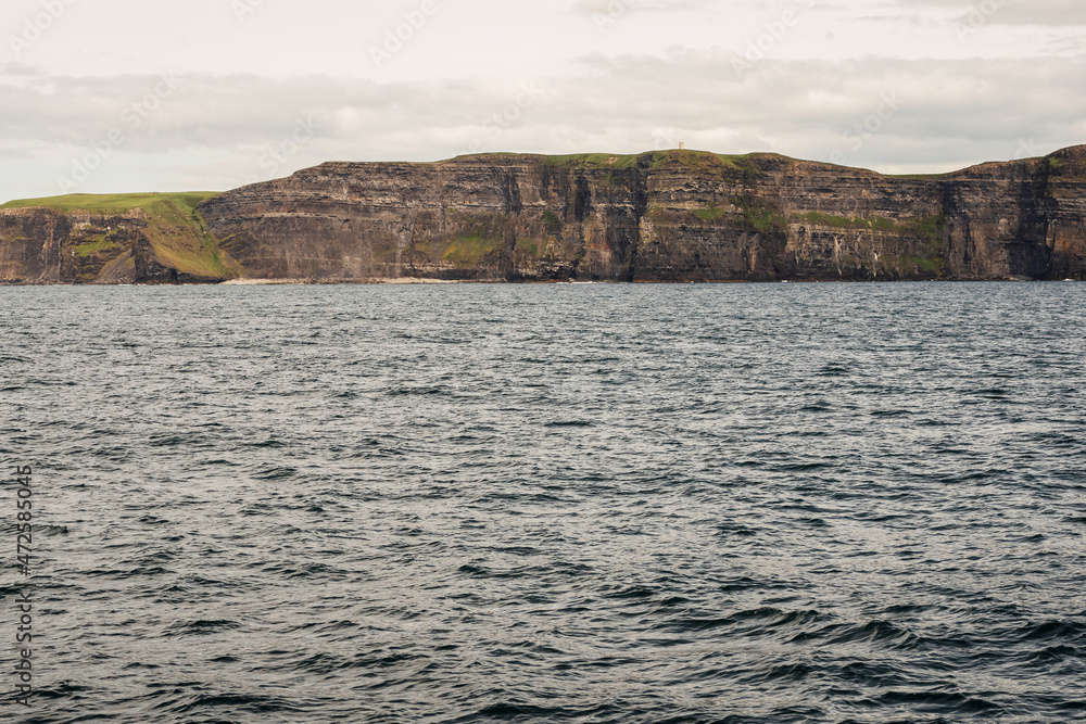 View on Cliff of Moher from a board of a ferry or cruise ship. West coast of Ireland. Popular tourist destination. National landmark. Cloudy sky. Calm surface of Atlantic ocean. View on Cliff of Moher from a board of a ferry or cruise ship. West coast of Ireland. Popular tourist destination. National landmark. Cloudy sky. Calm surface of Atlantic ocean.