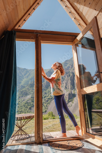 Happy woman on the porch of a small wooden house in the mountains. Concept of glamping and idyllic holidays outdoors © EdNurg