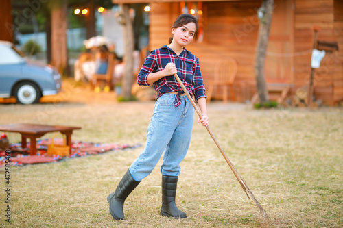 Fotografie, Obraz Young asian woman gardener in red plaid shirt smiling woman wearing boots black and holding rake with pile of autumn eaves and hay stack