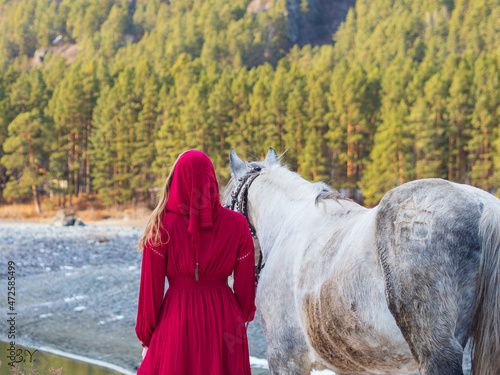 A girl in a red dress with a horse stands with her back to the viewer against the background of nature, an image from a fairy tale