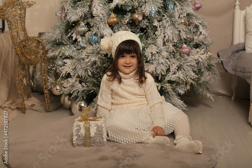 a little girl in a winter knitted hat and a white dress is sitting at the Christmas tree on a rug next to a gift box