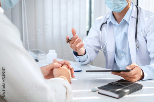 Male doctor in medical face mask is holding tablet to explaining about symptoms  treatment and giving advice to patient