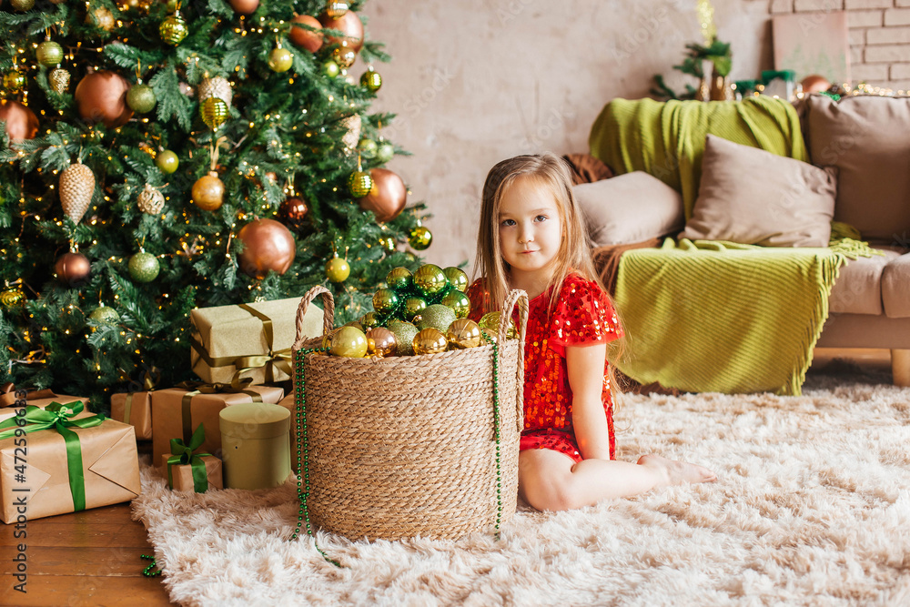 Little girl in a red dress with a Christmas tree toy in her hands sits at the Christmas tree. Christmas Holidays. Christmas decor, atmosphere, winter background