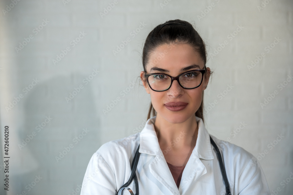 Portrait of young woman doctor standing in uniform with eyeglasses and stethoscope in the hospital office. Female health care medical worker in white coat looking at camera at the clinic room. Portrait of young woman doctor standing in uniform with eyeglasses and stethoscope in the hospital office. Female health care medical worker in white coat looking at camera at the clinic room.