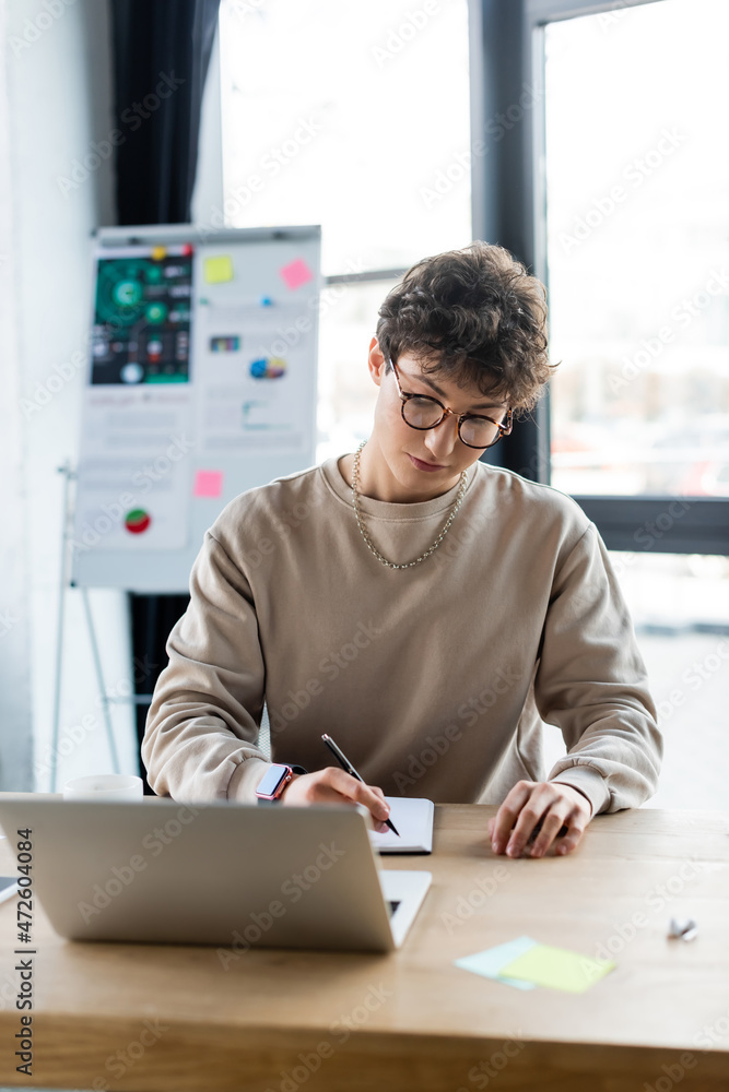 Young businessman writing on notebook near laptop and sticky notes in office.