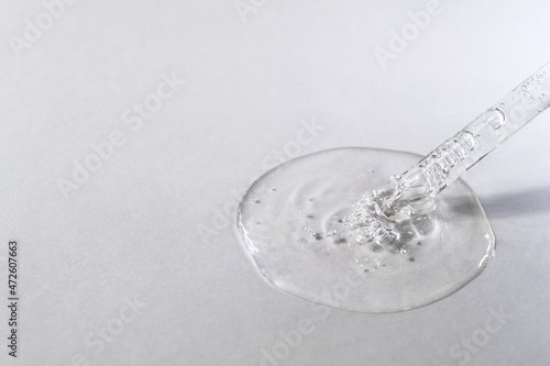 Transparent facial serum pouring from glass pipette on grey textured background. Liquid gel drop with bubbles closeup. Skin care routine. Trendy cosmetic product macro. Copy space, front view.