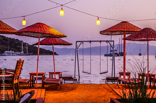 Beach umbrellas, sun loungers and swings under red lanterns against the backdrop of the sea, islands, mountains and yachts at sunset. Summer vacation concept