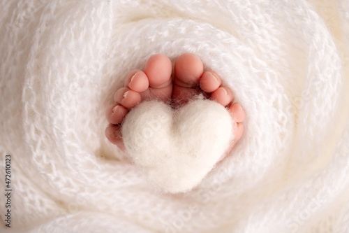 The tiny foot of a newborn baby. Soft feet of a new born in a white wool blanket. Close up of toes, heels and feet of a newborn. Knitted white heart in the legs of a baby. Studio macro photography. © Vad-Len