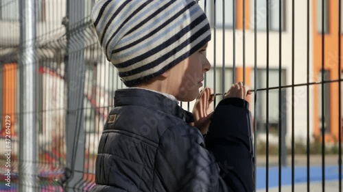 Upset and stressed boy looking through school metal fence after was bullied. Concept of poverty, immigration, bullying and kids stress