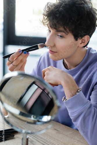 Young transgender person applying makeup foundation near mirror in studio.