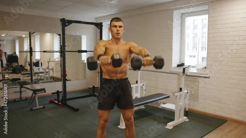 Young muscular man training in the gym and raising straight arms with dumbbells in front of him to work out the deltoid muscles