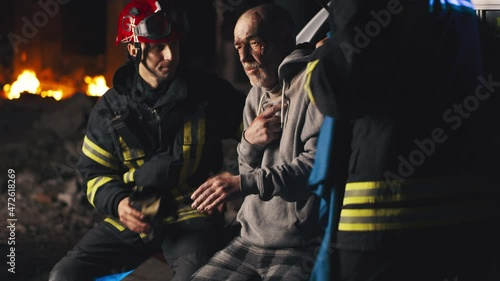 Male rescuers talking and putting blanket on shoulders of elderly man while paramedic wrapping bandage around injured hand of patient during work on disaster site at night photo