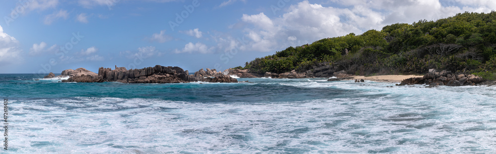 A high-resolution panorama of an waving oceanic landscape with huge stone formations in the middle. Turquoise water washes over the white sand beach. A rainforest of palm trees grows along the shore.