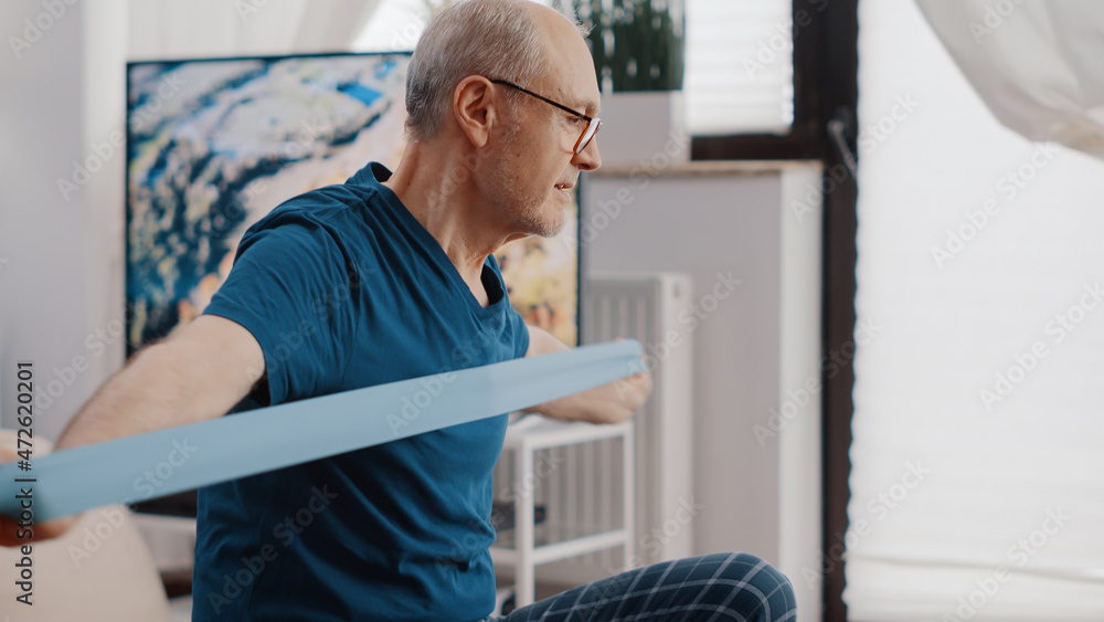 Close up of aged person pulling resistance band to exercise and sitting on fitness toning ball. Senior man training with elastic belt and looking at video of workout lesson on laptop