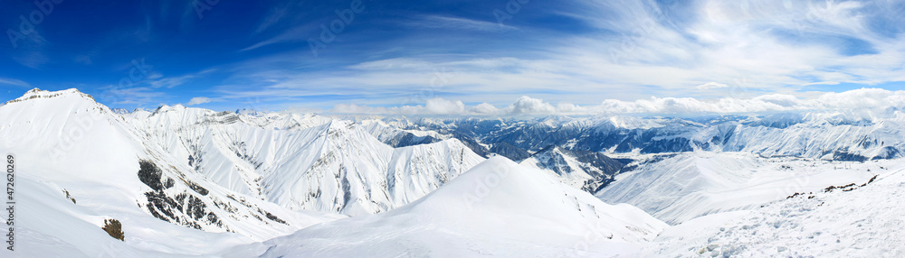 A view of the snowy mountains over which the clouds are floating. In the background is a blue sky with beautiful clouds. Panoramic photo.