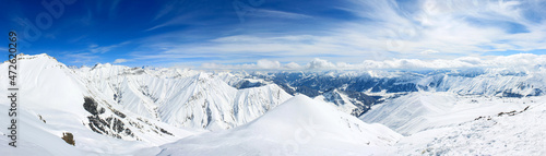 A view of the snowy mountains over which the clouds are floating. In the background is a blue sky with beautiful clouds. Panoramic photo.