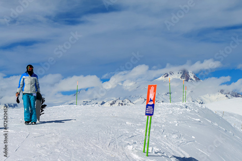 A girl with a snowboard in her hand stands alone on a ski slope. In front of her is a sign that reads "Avalanche Danger. In the background is a blue sky and mountain peaks in the clouds.