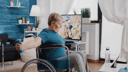 Woman with physical disability doing exercise with dumbbells and watching workout video on tablet. Retired person sitting in wheelchair and following training lesson with weights.
