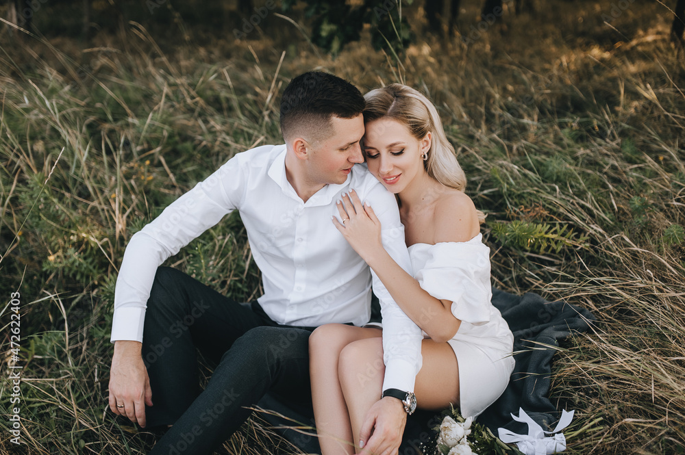 Stylish groom and cute blonde bride with curly hair in a white dress are sitting on a rug on the yellow grass in nature and hugging. Wedding portrait of beautiful newlyweds. Stylish groom and cute blonde bride with curly hair in a white dress are sitting on a rug on the yellow grass in nature and hugging. Wedding portrait of beautiful newlyweds.