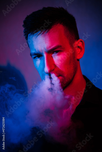 a young man in a neon light with a stern look blows a lot of smoke from his nose