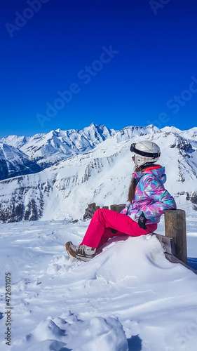 Snowboarder girl sitting on the side of a slope admiring the tall Alps. Slopes are covered with powder snow. Perfect weather for a ride. High alpine snowboarding. Girl wears a helmet for protection