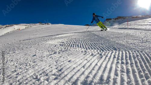A skier going down the slope in Heiligenblut, Austria. Perfectly groomed slopes. High mountains surrounding the man wearing yellow trousers and blue jacket. Man wears helm for the protection.