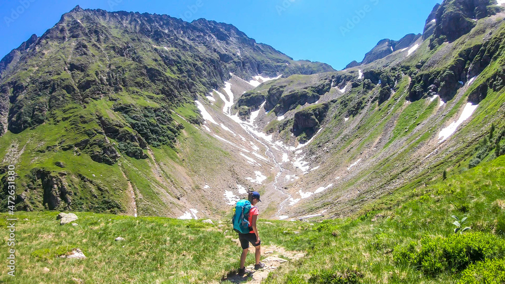 Obraz premium A young woman with a big backpack hikes down on a steep pathway between tall mountain peaks. Some of the slopes are covered with snow. In the back is another mountain range. Spring in alpine valleys.