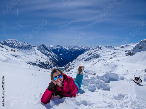 A girl in a snowboarding outfit lies on the snow and enjoys her break. Endless chains of snow caped Alps surrounding the girl. Girl is smiling. Clear and bright day. Moelltaler Gletscher, Austria.