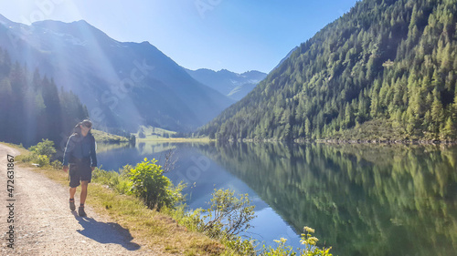 Young man walking along an alpine lake. The lake is surrounded with tall mountains. The surface of the lake is calm, it reflects the mountains and sky. Clear and sunny day. Schladming region, Austria photo