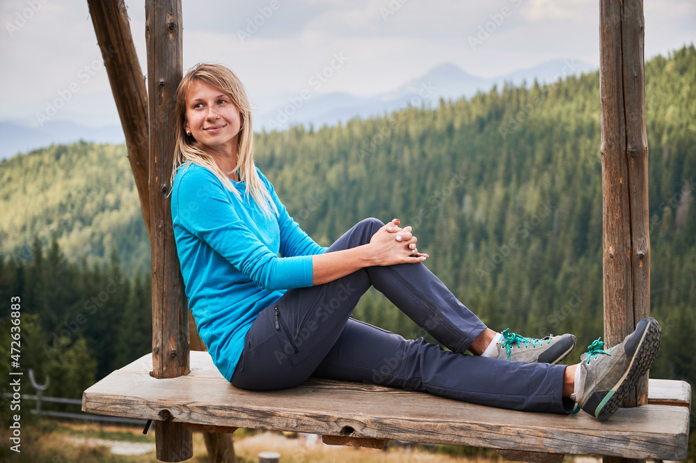 Nice girl resting on wooden seat suspended on two wooden poles and enjoying amazing scenery. On the background green forest and silhouettes of mountain hills far away. Nice girl resting on wooden seat suspended on two wooden poles and enjoying amazing scenery. On the background green forest and silhouettes of mountain hills far away.
