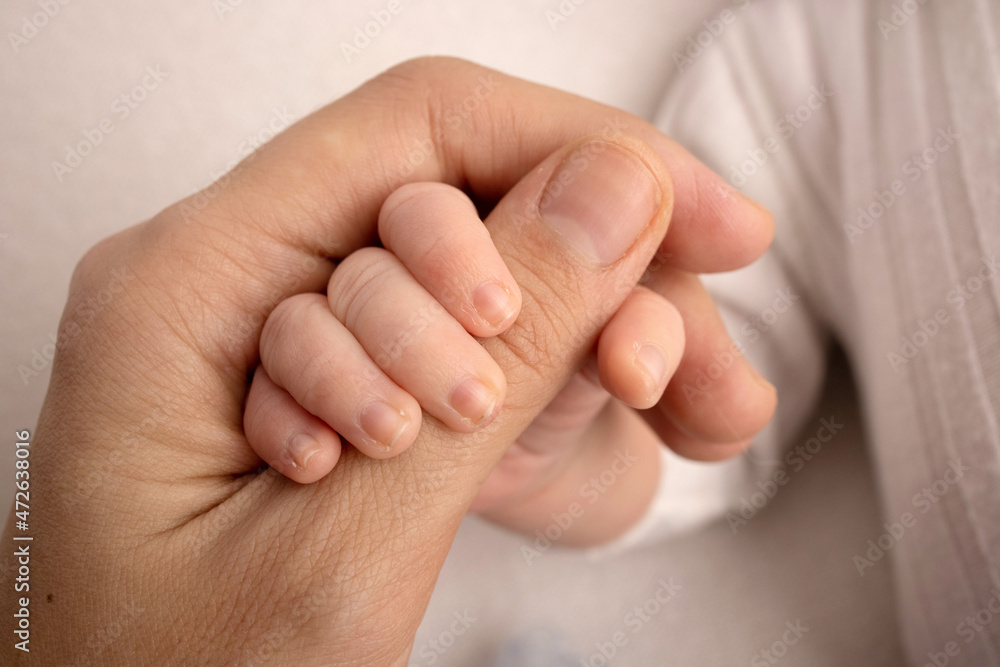 The newborn baby in a white closes has a firm grip on the parent's finger after birth. Close-up little hand of child and palm of mother and father. Parenting, childcare and healthcare concept. The newborn baby in a white closes has a firm grip on the parent's finger after birth. Close-up little hand of child and palm of mother and father. Parenting, childcare and healthcare concept.