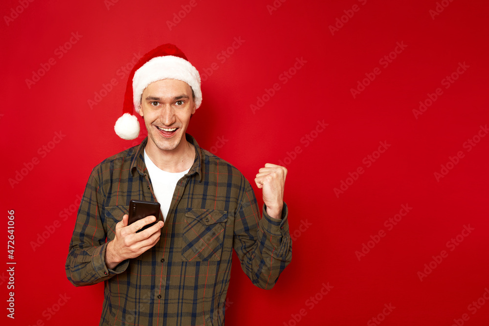 concept - people, technology, success. an excited joyful man with phone in hand, New Year hat, celebrates victory in mobile application game, received good news. isolated on red studio background concept - people, technology, success. an excited joyful man with phone in hand, New Year hat, celebrates victory in mobile application game, received good news. isolated on red studio background