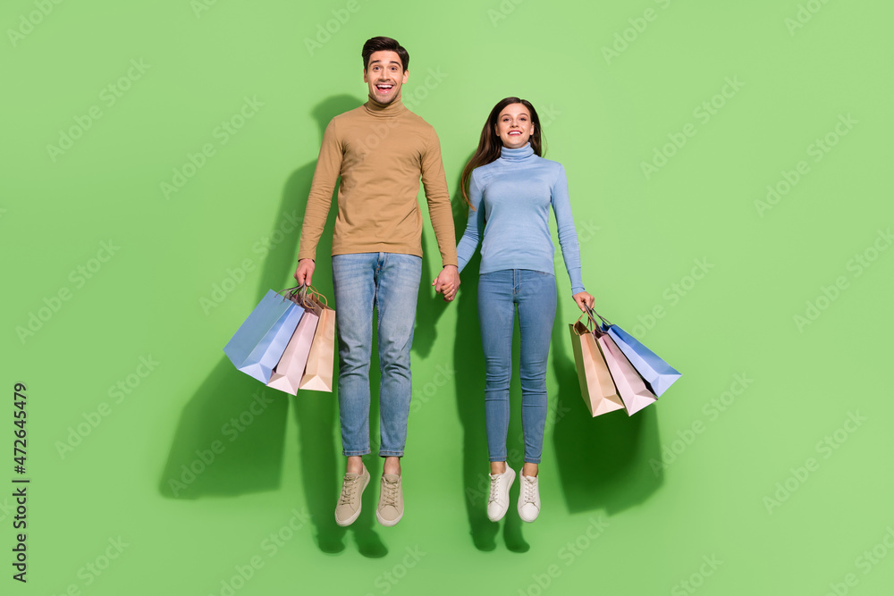 Full length portrait of two overjoyed cheerful persons hold arms packages isolated on green color background Full length portrait of two overjoyed cheerful persons hold arms packages isolated on green color background