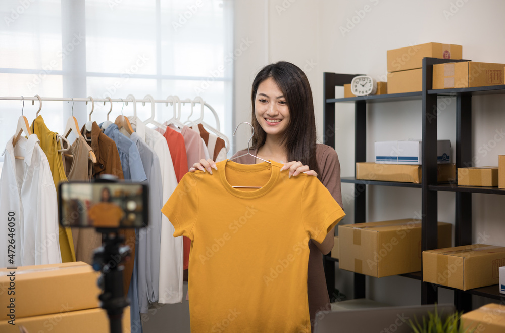 Young asian woman business owner at fashion store using smartphone live streaming for sale fashion clothing. Blogger record video review clothes for sell on social media. Online shopping concept. Phot...