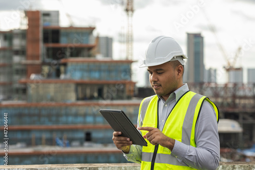 Asian engineer handsome man or architect use tablet with white safety helmet in city construction site . Standing on rooftop building construction at capital.