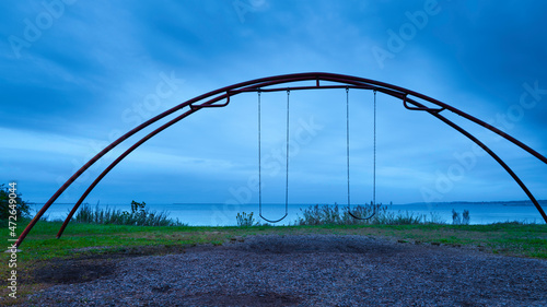 Dramatic twilight seascape with a swing set and the arching twin metal frames in the beach park over the green. Empty tranquil off-season coastal feature in New Haven Harbor, Connecticut.