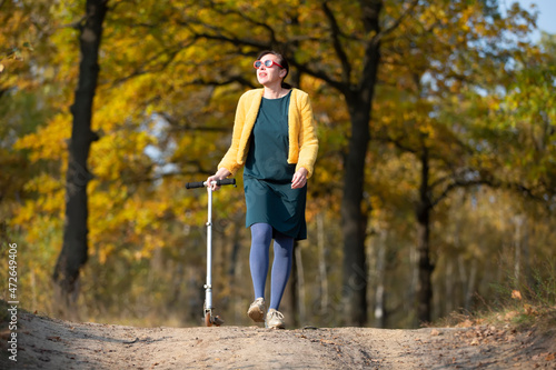 A beautiful middle-aged woman walks with a scooter in autumn.