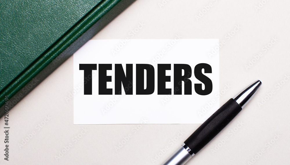 On a light gray background lies a pen, a green notebook and a white card with the text TENDERS. Business concept. On a light gray background lies a pen, a green notebook and a white card with the text TENDERS. Business concept.