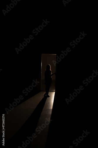 A silhouette of a little girl emerges from a luminous room with smoke in the dark.