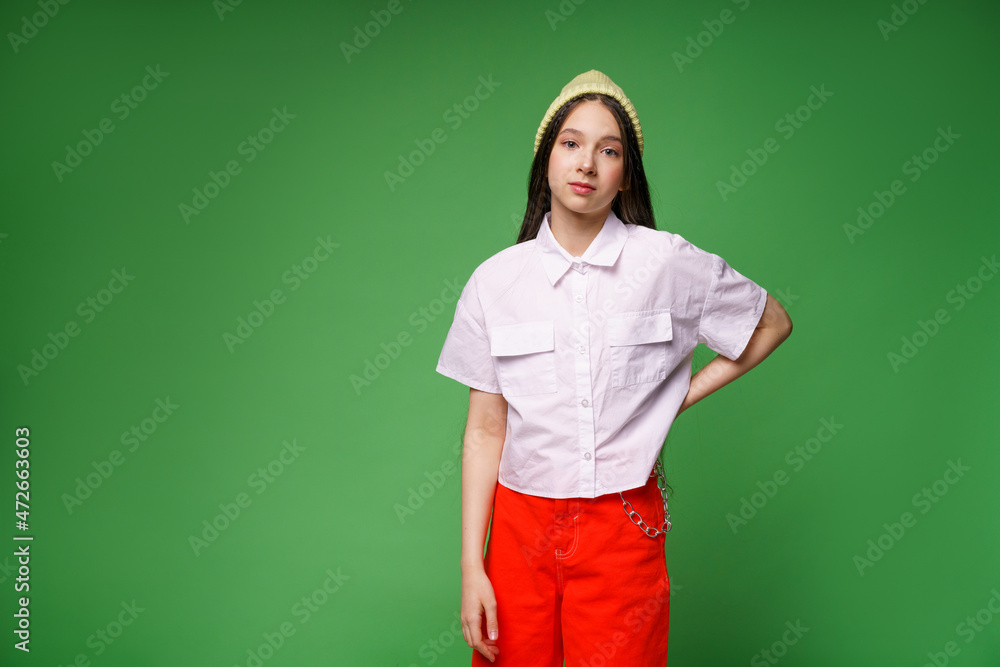 Beautiful cute millennial girl in hat and white shirt and in red pants isolated on green studio background, happy young woman in looking at camera, posing or casting