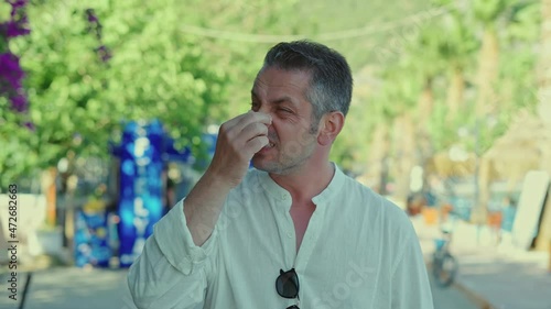 Young bearded man coughing while walking outside, feeling sick.Man with pollen allergy running tears, eyes itching and sneezing on a beautiful flowery road. The concept of living with allergies.