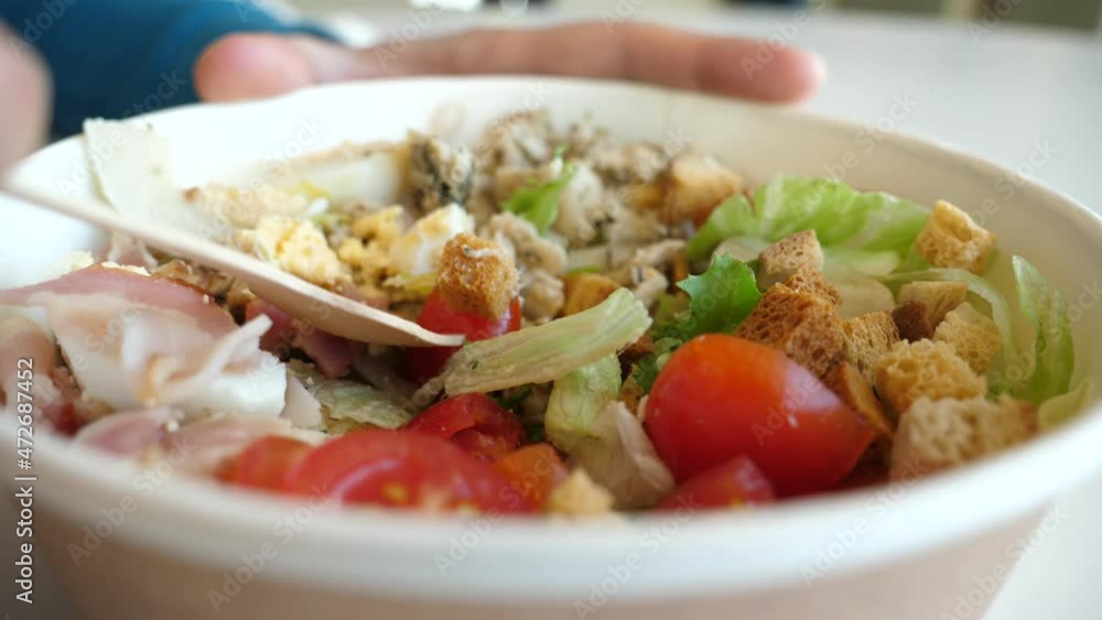 Man eating a Caesar salad of vegetables and beacon meat, eggs and croutons, red tomatoes, from disposable paper bowl use wooden fork in food court of shopping center Man eating a Caesar salad of vegetables and beacon meat, eggs and croutons, red tomatoes, from disposable paper bowl use wooden fork in food court of shopping center