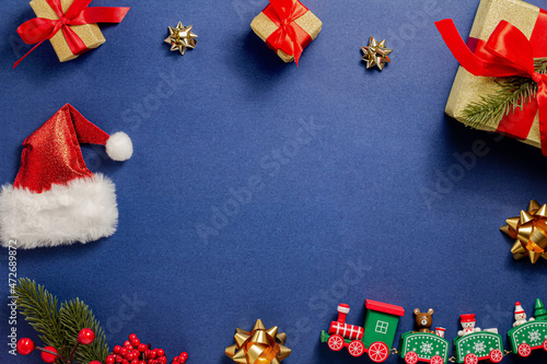 Christmas composition. A wooden train  a Christmas tree with red berries  golden bows  gifts  a Santa hat on a dark blue background. New Year. Flat lay  top view  copy space