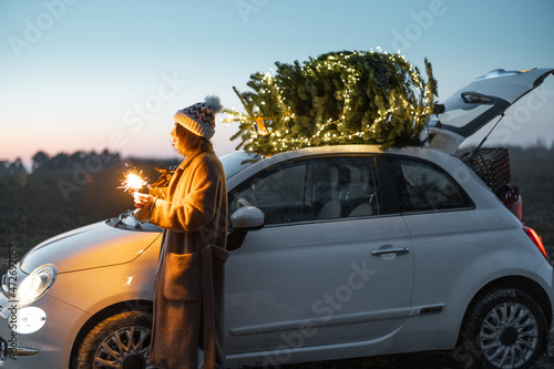 Fototapeta Naklejka Na Ścianę i Meble -  Happy caucasian woman firing sparklers near car with Christmas tree on nature at dusk. Concept of celebrating New Year holidays. Idea of Christmas mood and fun. Side view with sunset sky on background