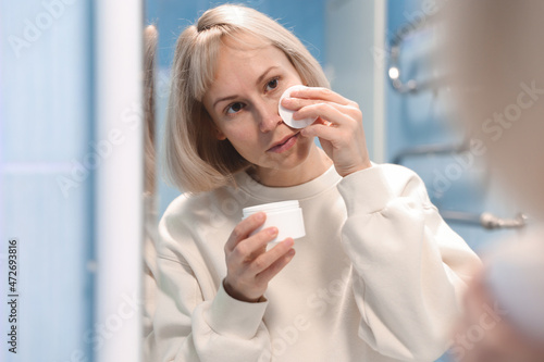 A woman reflected in the bathroom mirror cleanses her face with a sponge for sensitive skin, gentle hygienic daily use, effective removal of impurities, skin care concept © Enigma